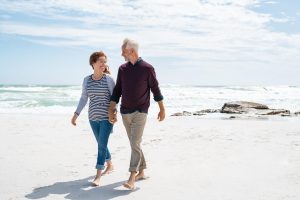 Senior couple walking and looking at each other at beach. Happy mature couple in love in a bright sunny day walking barefoot at sea shore. Loving retired man with beautiful casual woman relaxing at the ocean with copy space.